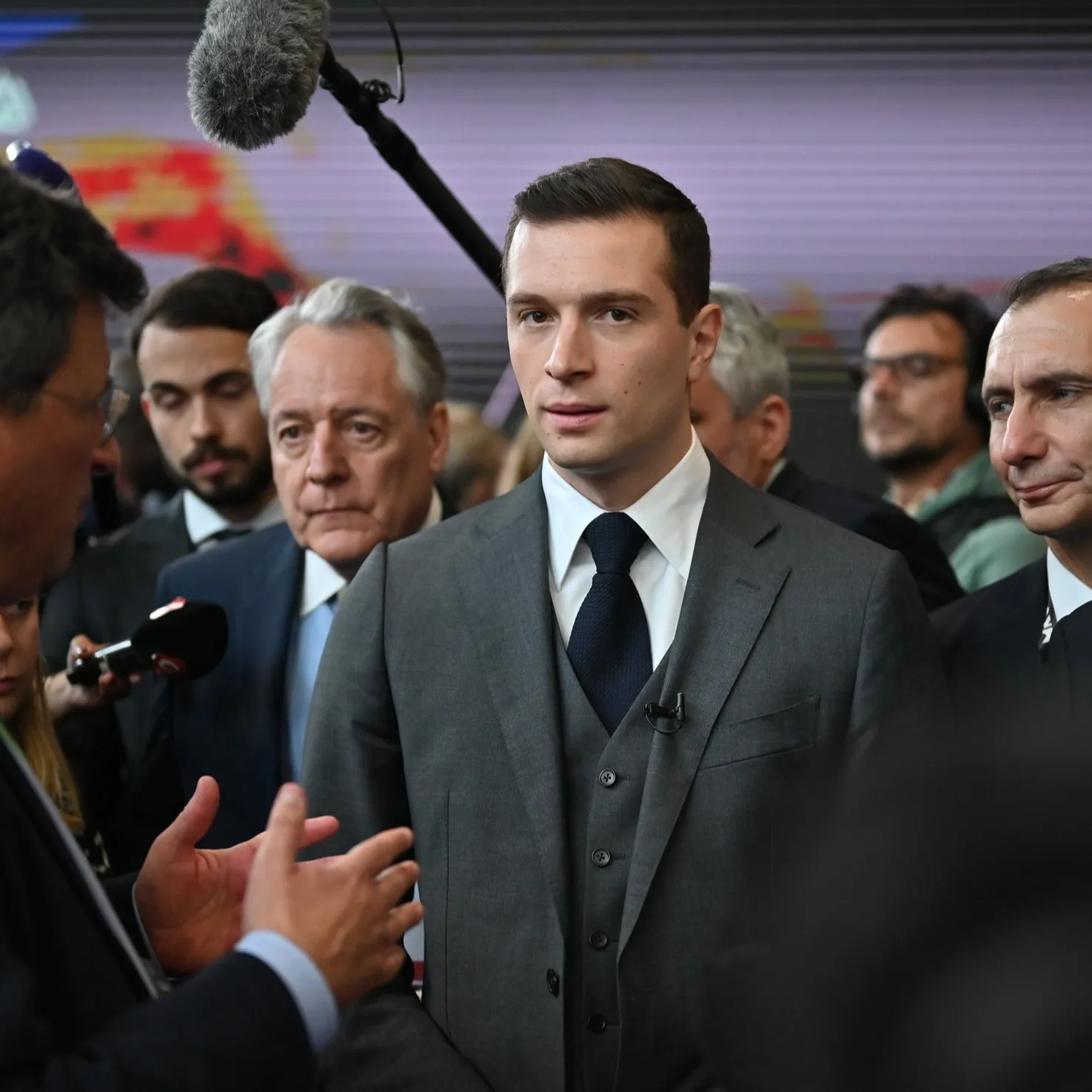 A man in a suit stands with a group of other men. There is a microphone above him