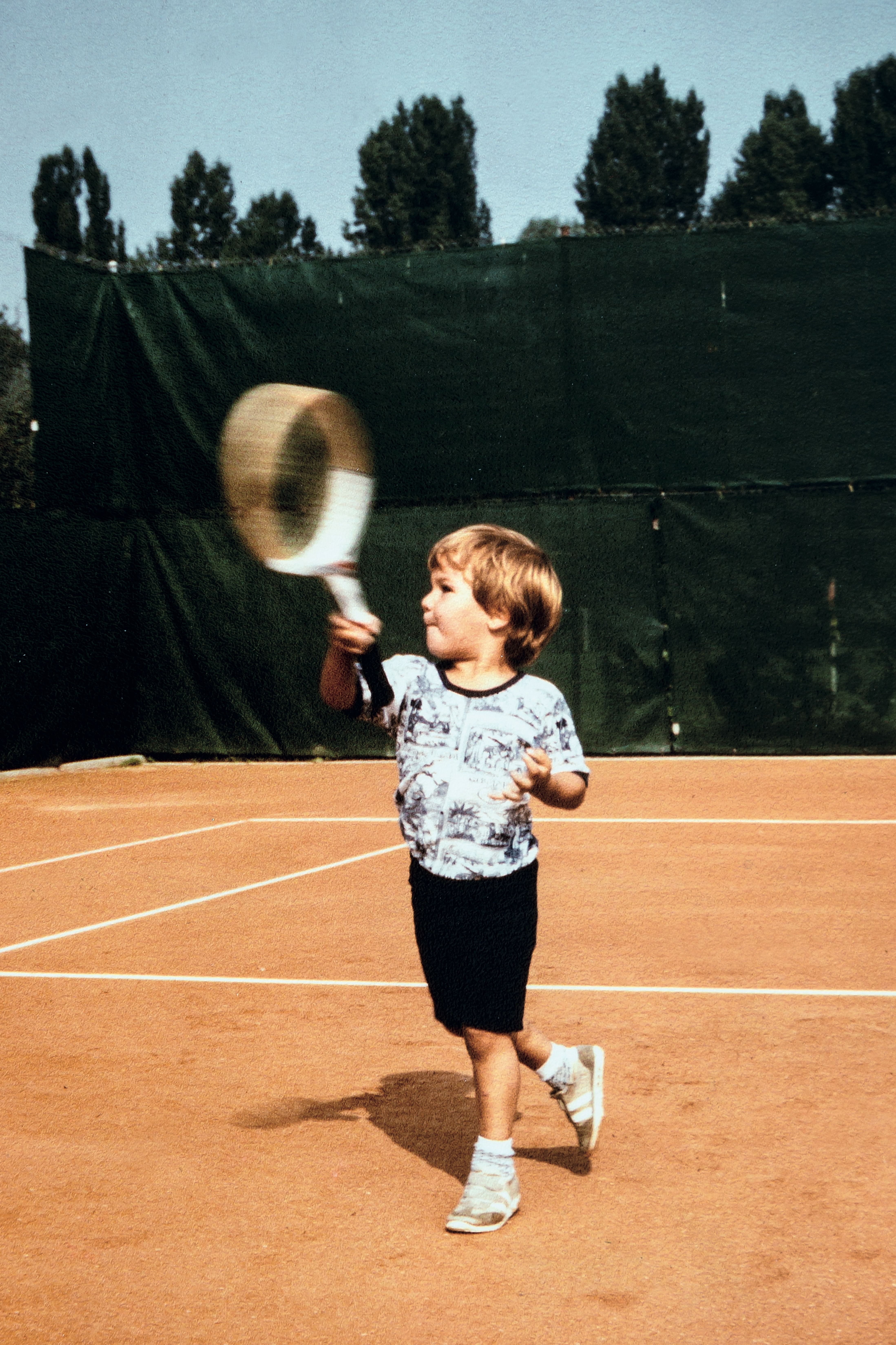 A young boy, 罗杰•费德勒, holds up a tennis racket as if to return a shot, on a clay court. The photo was taken in Basel, Switzerland, where费德勒was born in 1981